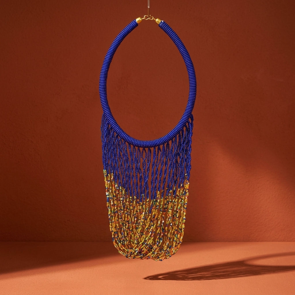 A large statement necklace with a thick blue beaded collar and long fringe that transitions into gold-toned beads, hanging against a warm terracotta background.