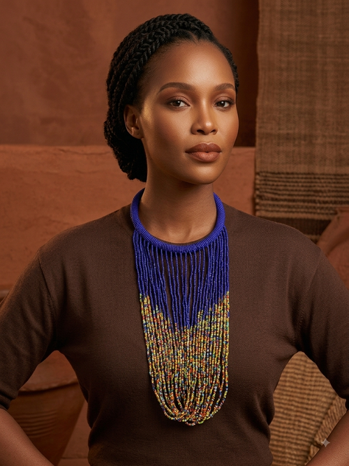 A woman wearing a brown top and a large beaded necklace with a royal blue top section and multicolor dangling strands, posed against a warm brown textured background.